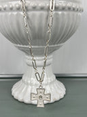 A silver-toned paperclip chain necklace with an elongated rectangular link design, featuring a decorative cross pendant with small pearl accents at the center, displayed on a white ribbed ceramic pedestal.