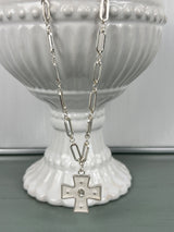 A silver-toned paperclip chain necklace with an elongated rectangular link design, featuring a decorative cross pendant with small pearl accents at the center, displayed on a white ribbed ceramic pedestal.
