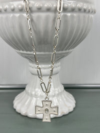 A silver-toned paperclip chain necklace with an elongated rectangular link design, featuring a decorative cross pendant with small pearl accents at the center, displayed on a white ribbed ceramic pedestal.
