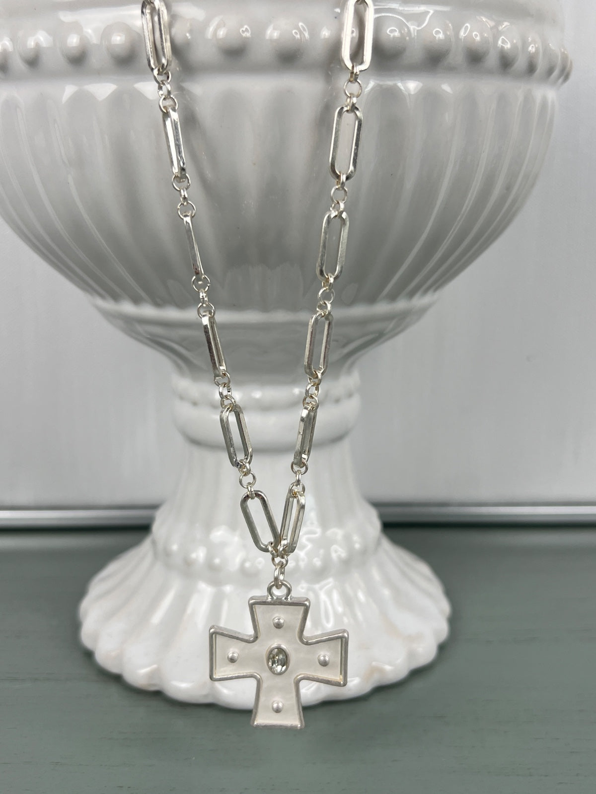 A silver-toned paperclip chain necklace with an elongated rectangular link design, featuring a decorative cross pendant with small pearl accents at the center, displayed on a white ribbed ceramic pedestal.