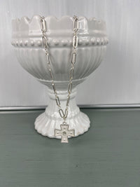 A silver-tone chain necklace with an elongated cross pendant featuring a small rhinestone accent, displayed on a white ribbed pedestal bowl against a soft green surface.
