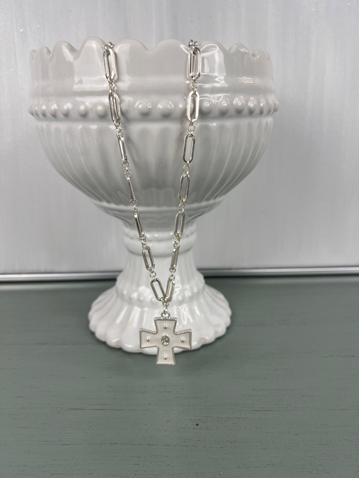 A silver-tone chain necklace with an elongated cross pendant featuring a small rhinestone accent, displayed on a white ribbed pedestal bowl against a soft green surface.