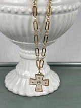 A gold-toned chain necklace with elongated oval links featuring a decorative cross pendant with studded details, displayed on a white ceramic pedestal vase against a soft green background.
