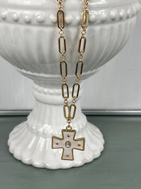 A gold-toned chain necklace with elongated oval links featuring a decorative cross pendant with studded details, displayed on a white ceramic pedestal vase against a soft green background.