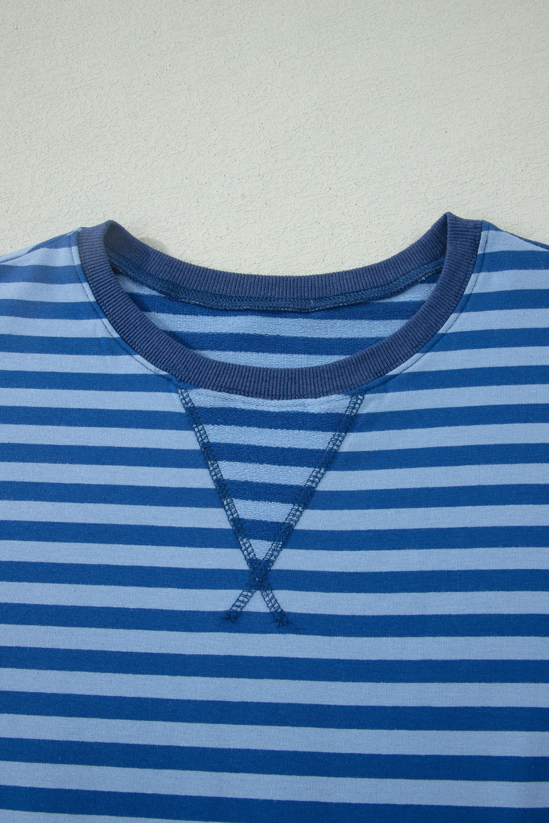 A close-up of a striped long-sleeve shirt featuring alternating navy and light blue horizontal stripes with a navy ribbed crew neckline and decorative cross-stitch detailing on the chest.