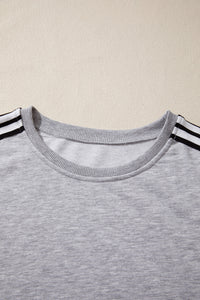 A light gray crewneck t-shirt with black and white striped trim on the short sleeves, displayed flat against a beige background.