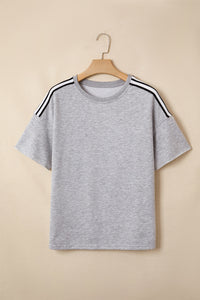 A light gray short-sleeve t-shirt on a wooden hanger featuring black and white striped trim along the shoulders. The shirt has a simple crew neckline and relaxed fit displayed against a neutral beige background.