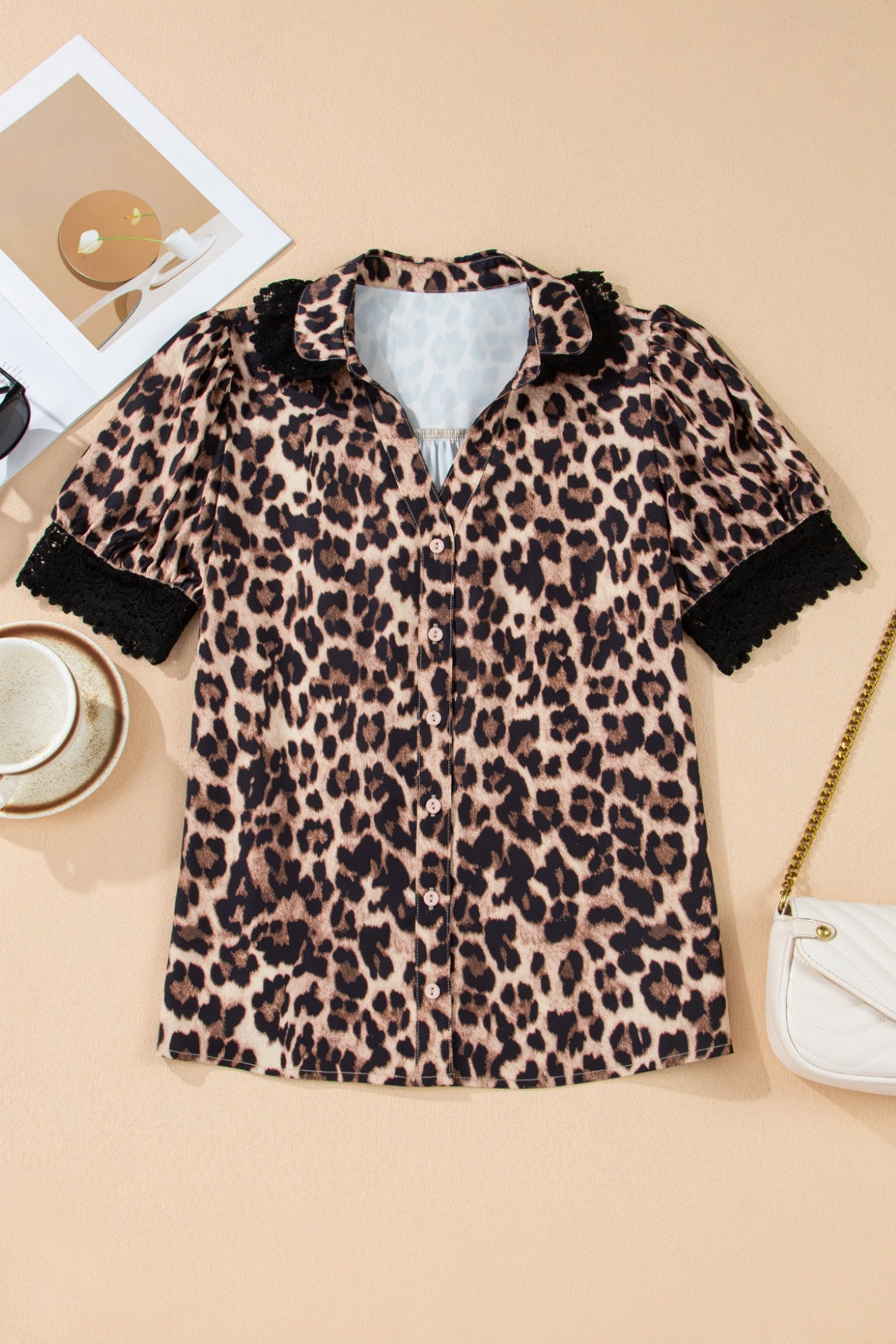 A leopard print short-sleeve shirt with a button-front closure and black ruffled collar and cuffs, displayed flat lay on a beige background alongside accessories including a white crossbody bag and sunglasses.