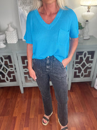 A woman wearing a bright turquoise ribbed short-sleeve V-neck sweater tucked into dark wash denim jeans, styled with white and black slide sandals.