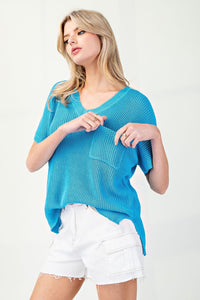 A woman wearing a bright turquoise short-sleeve V-neck ribbed knit top with an asymmetrical hem, paired with white distressed denim shorts.