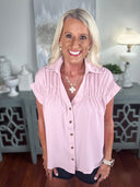A woman wearing a light pink short-sleeve button-up blouse with ruched detailing at the shoulders, paired with dark shorts. The blouse features a collared neckline and is styled with a silver cross necklace and watch.