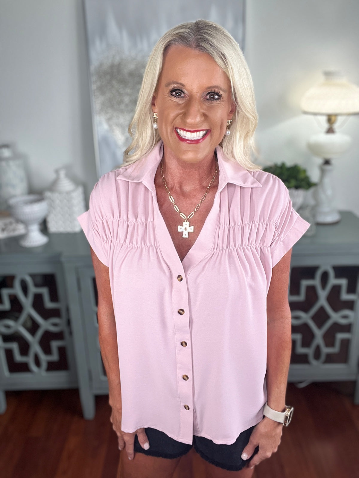 A woman wearing a light pink short-sleeve button-up blouse with ruched detailing at the shoulders, paired with dark shorts. The blouse features a collared neckline and is styled with a silver cross necklace and watch.
