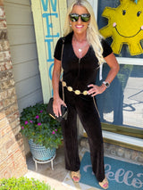 A woman wearing a black pinstriped short-sleeve jumpsuit with a gold chain belt, paired with mirrored sunglasses, a black crossbody bag, and tan sandals, standing in front of a storefront.