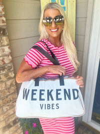 A woman wearing a hot pink and white striped short-sleeve t-shirt holds a white canvas tote bag with black handles that reads "WEEKEND VIBES" in dark text. She accessorizes with mirrored sunglasses and a crossbody bag while smiling at the camera.
