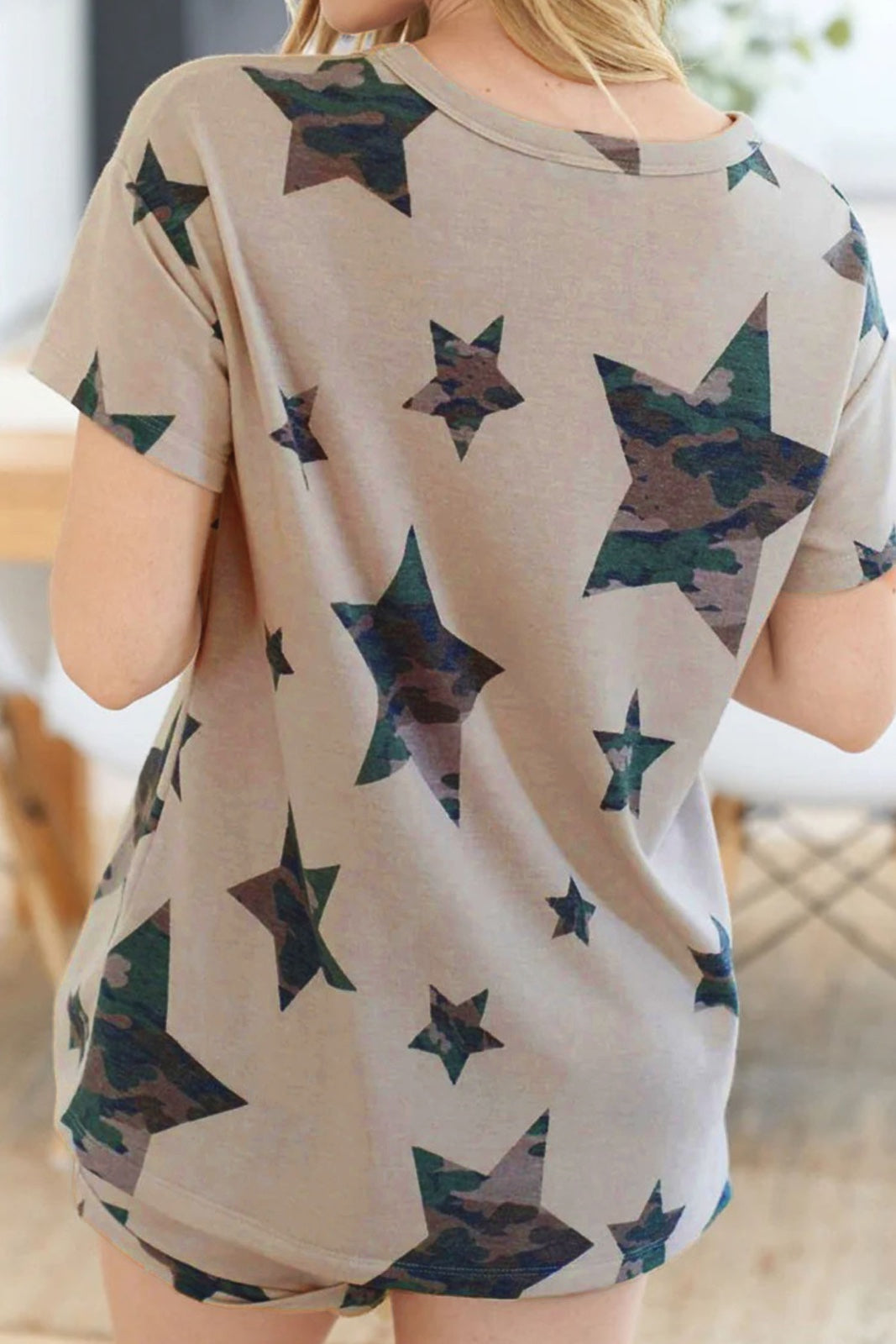 Woman wearing a beige short-sleeve t-shirt with an all-over print of dark teal and brown camouflage-patterned stars in varying sizes, photographed from the back.