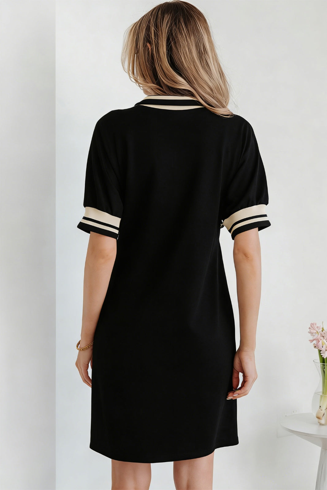 A woman viewed from behind wearing a black short-sleeve dress with cream and black striped trim at the neckline and cuffs, featuring a simple A-line silhouette.