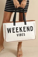 A person wearing a black and white striped dress holds a large cream canvas tote bag with "WEEKEND VIBES" text printed in black and tan leather trim, featuring black handles.