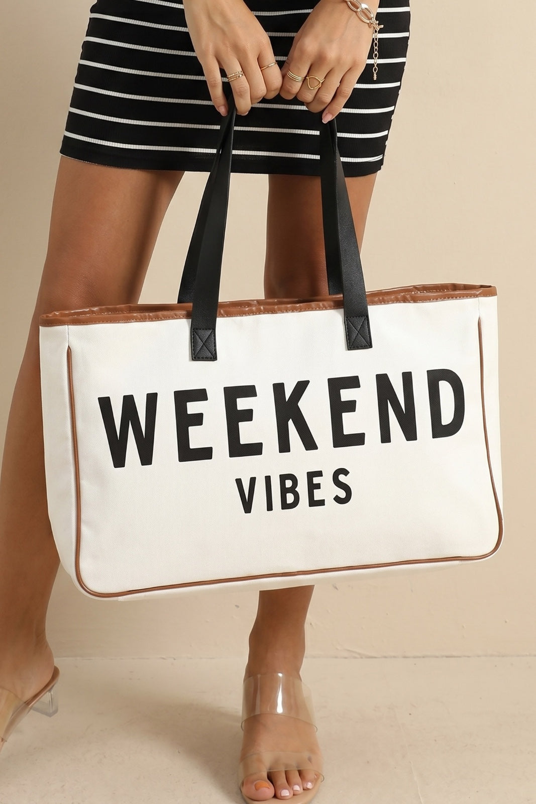 A person wearing a black and white striped dress holds a large cream canvas tote bag with "WEEKEND VIBES" text printed in black and tan leather trim, featuring black handles.