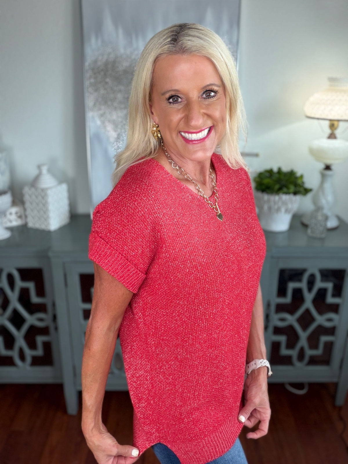 Woman wearing a bright red short-sleeved knit sweater with a textured fabric, paired with jeans, photographed in an indoor home setting.