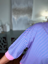 A blue and white striped long-sleeve shirt with pink cuffs, displayed on a person's shoulder and arm in an indoor setting.
