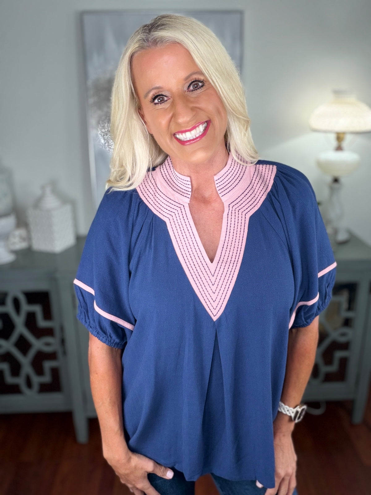 A woman wearing a navy blue short-sleeved tunic top with pink and white striped V-neckline trim and matching striped cuffs, paired with dark jeans.