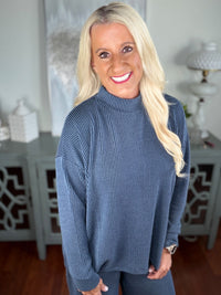 A woman wearing a blue ribbed knit sweater with a crew neckline and long sleeves, styled with jeans in a bright, neutral indoor setting.