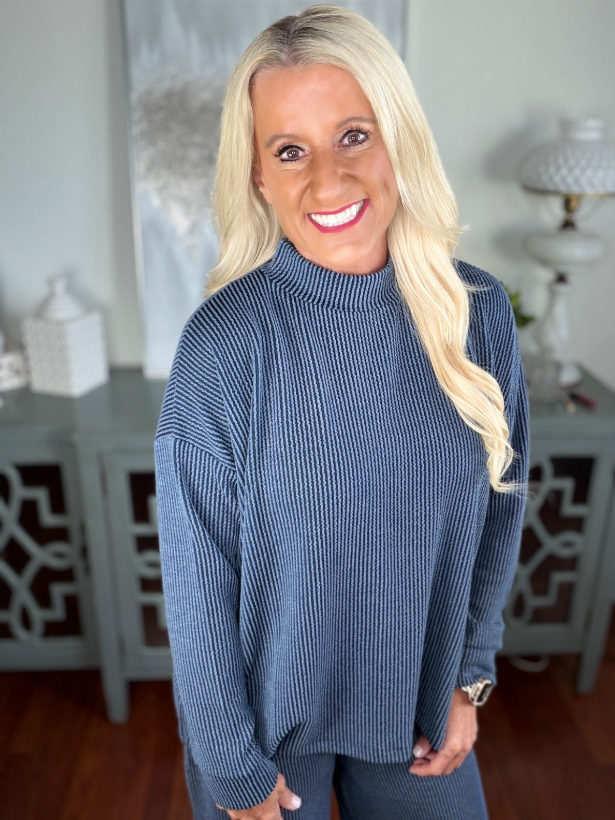 A woman wearing a blue ribbed knit sweater with a crew neckline and long sleeves, styled with jeans in a bright, neutral indoor setting.