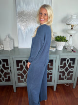 Woman wearing a long blue ribbed cardigan with long sleeves over matching pants, styled casually while posing next to a decorative teal cabinet.