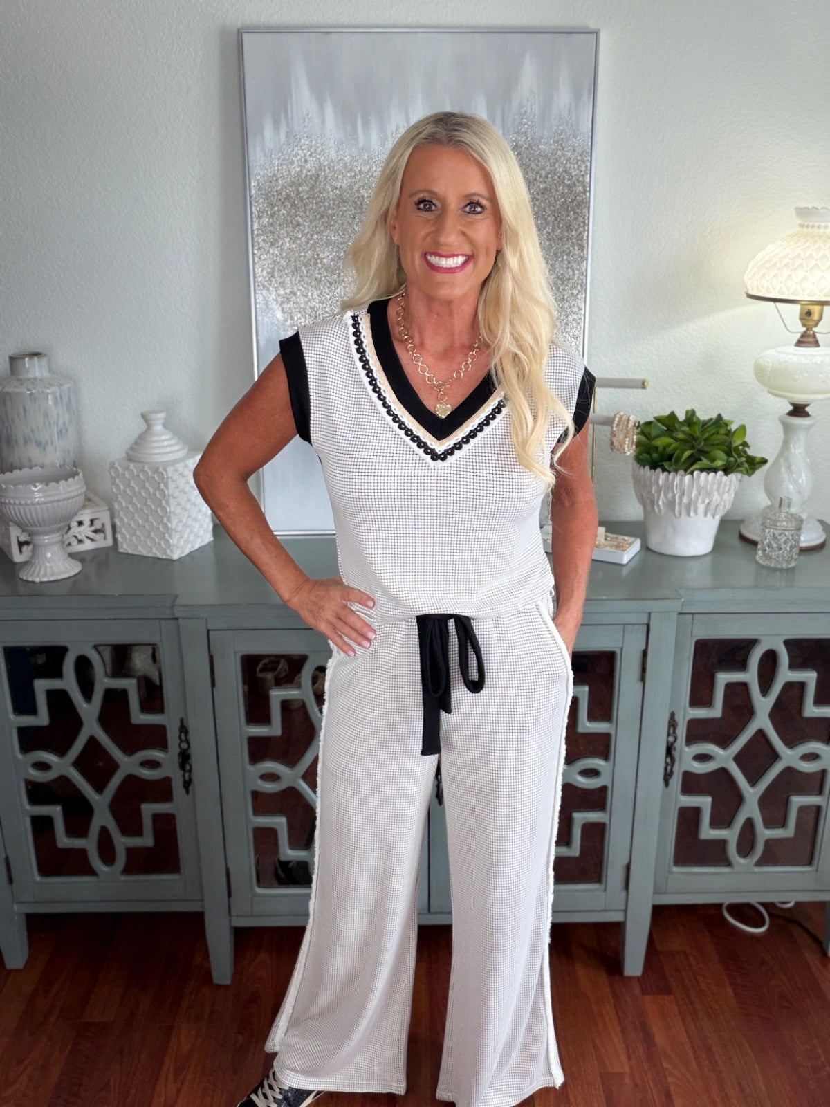 A woman wearing a sleeveless white ribbed knit jumpsuit with black and white V-neckline trim and a black drawstring waist, paired with a gold chain necklace.