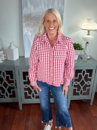 A woman wearing a red and white gingham button-up shirt with ruffled trim details, paired with distressed blue denim jeans and white sneakers, standing in front of a gray cabinet with decorative accessories.
