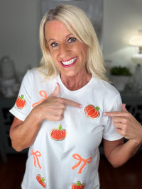A woman wearing a white short-sleeved t-shirt decorated with red apple prints and orange bow designs throughout. She is smiling at the camera and pointing to the shirt's pattern.