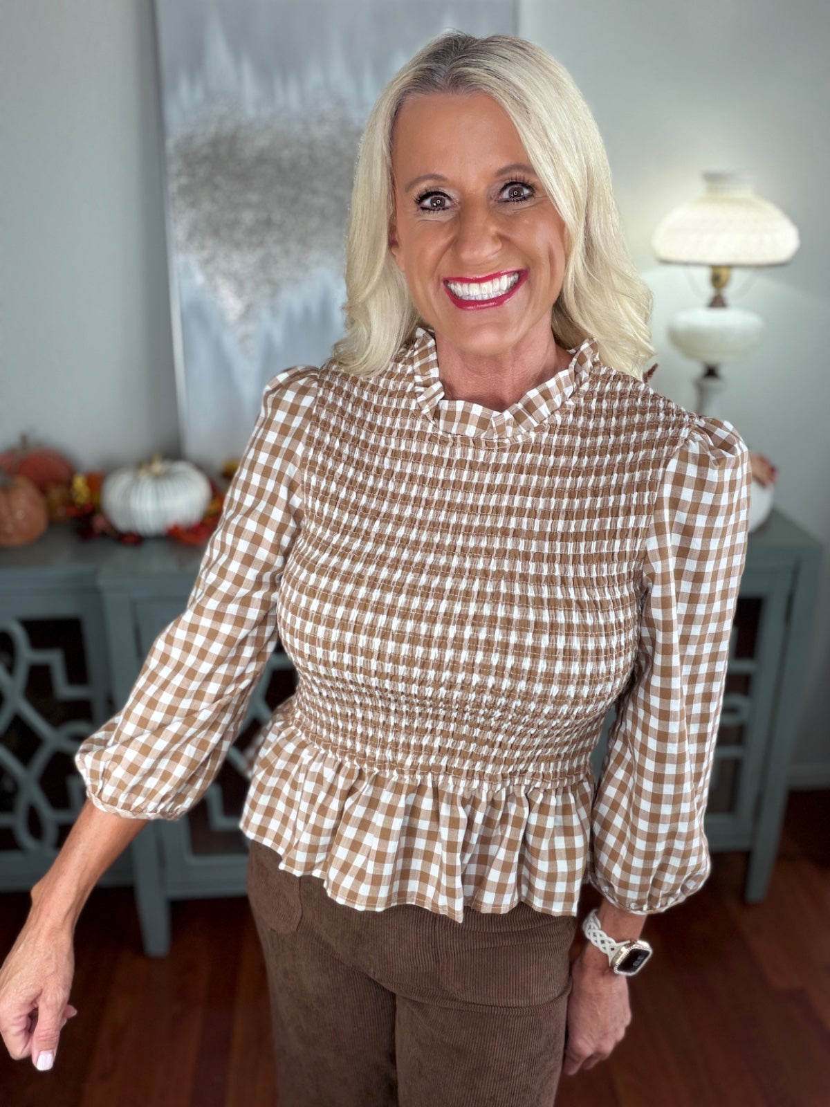 Woman wearing a brown and white gingham check blouse with three-quarter length sleeves and a smocked peplum waist, paired with brown ribbed knit pants.