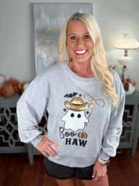 A woman wearing a light gray sweatshirt featuring a cheerful ghost character wearing a cowboy hat with "Boo HAW" text and Halloween-themed graphics in black and gold embroidery.