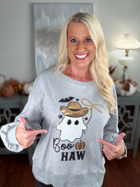A woman wearing a light gray crewneck sweatshirt featuring a cute ghost design with a straw hat, bat, and "Boo HAW" text in black and tan colors. The sweatshirt has ribbed cuffs and is styled with a black shorts and white sneakers.