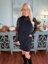 Woman wearing a black long-sleeve dress with lace sleeves and a ribbed knit body, paired with black knee-high boots, standing in front of an autumn-decorated console table.