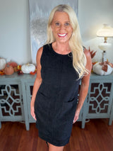 A woman wearing a sleeveless black ribbed knit dress that falls to knee length, styled with a simple crew neckline. The dress is photographed in an interior setting decorated with autumn-themed accessories.