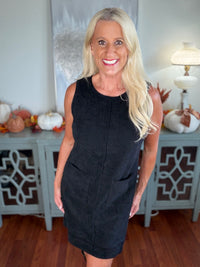 A woman wearing a sleeveless black ribbed knit dress that falls to knee length, styled with a simple crew neckline. The dress is photographed in an interior setting decorated with autumn-themed accessories.
