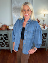 A woman wearing an oversized medium-wash denim shirt with button front, chest pocket, and rolled sleeves over a black top and brown pants, styled in a casual, relaxed fit.