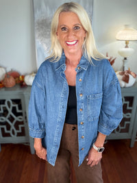 A woman wears an oversized denim shirt jacket in light blue wash, featuring a classic collar, button front closure, and chest pocket, styled over a black top with brown corduroy pants.