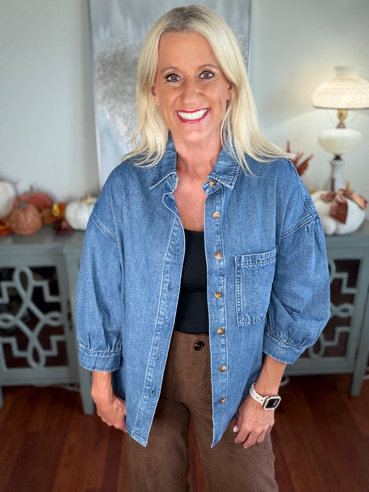 A woman wears an oversized denim shirt jacket in light blue wash, featuring a classic collar, button front closure, and chest pocket, styled over a black top with brown corduroy pants.