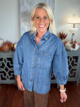 A woman wears an oversized denim shirt in medium blue wash, featuring a classic collar, button front with copper-tone buttons, and a chest pocket. The shirt has three-quarter length sleeves and is styled casually over brown pants.