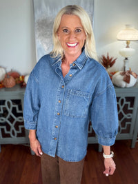 A woman wears an oversized denim shirt in medium blue wash, featuring a classic collar, button front with copper-tone buttons, and a chest pocket. The shirt has three-quarter length sleeves and is styled casually over brown pants.