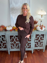 A woman wearing a burgundy waffle-knit long-sleeve sweater paired with matching burgundy leggings, styled with white sneakers against a fall-decorated sideboard backdrop.