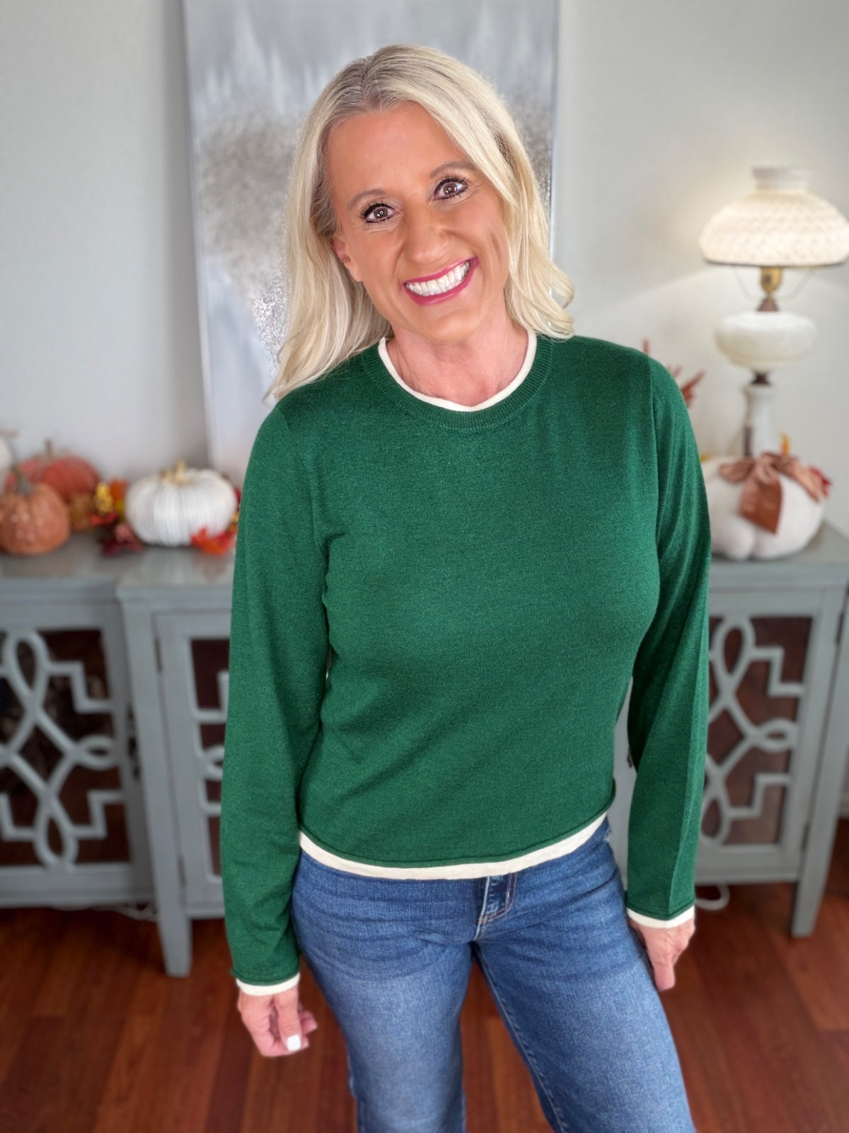 A woman wearing a forest green long-sleeve crewneck sweater with white ribbed trim at the neckline, cuffs, and hem, paired with blue jeans.