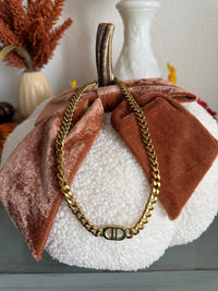A decorative fabric pumpkin with white textured base and rust-brown velvet leaves, featuring a brown wooden stem and a gold chain necklace with a geometric pendant draped across it.