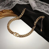 A gold-toned chunky curb chain bracelet featuring a decorative oval pendant in the center, displayed on a black jewelry stand next to a gold-framed mirror and white lace fabric.