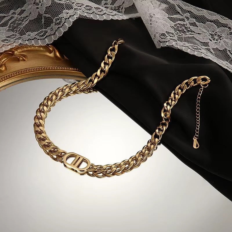 A gold-toned chunky curb chain bracelet featuring a decorative oval pendant in the center, displayed on a black jewelry stand next to a gold-framed mirror and white lace fabric.