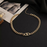 A gold-toned chunky chain necklace featuring a prominent DD pendant charm, displayed on a black surface alongside fashion magazines and a perfume bottle.