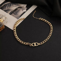 A gold-toned chunky chain necklace featuring a prominent DD pendant charm, displayed on a black surface alongside fashion magazines and a perfume bottle.
