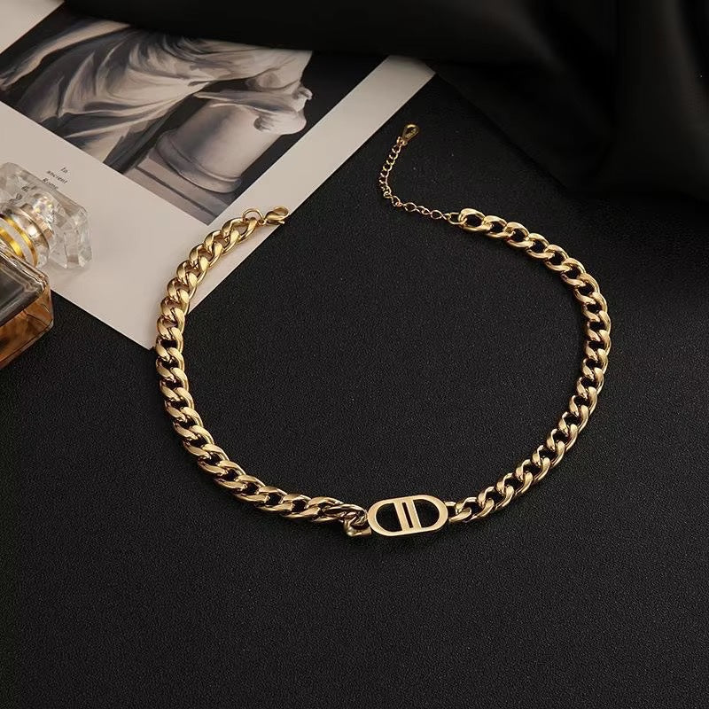 A gold-toned chunky chain necklace featuring a prominent DD pendant charm, displayed on a black surface alongside fashion magazines and a perfume bottle.